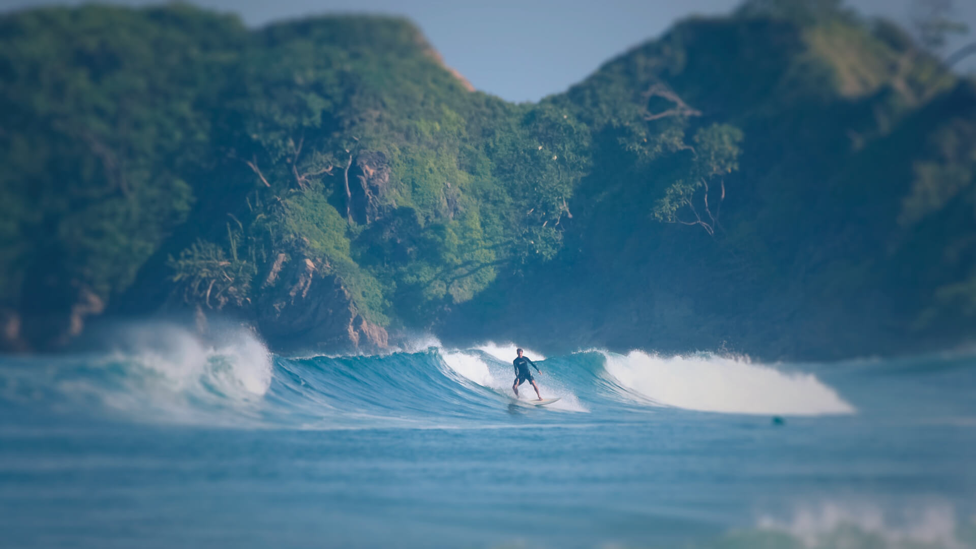 Surfer riding a wave with lush green hills in the background at Playa Guiones, Nosara