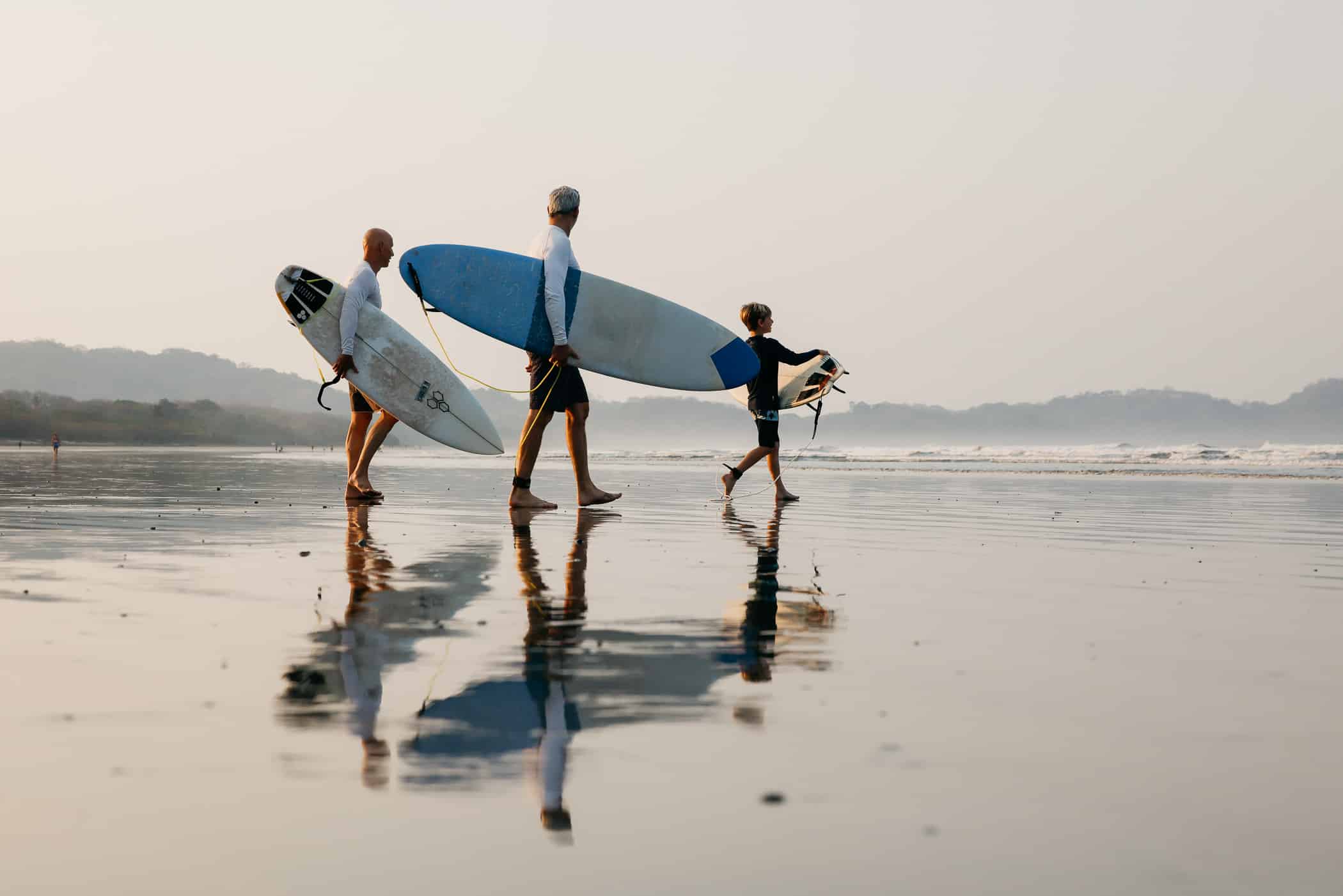 Family carrying surfboards across the reflective wet sand at Playa Guiones