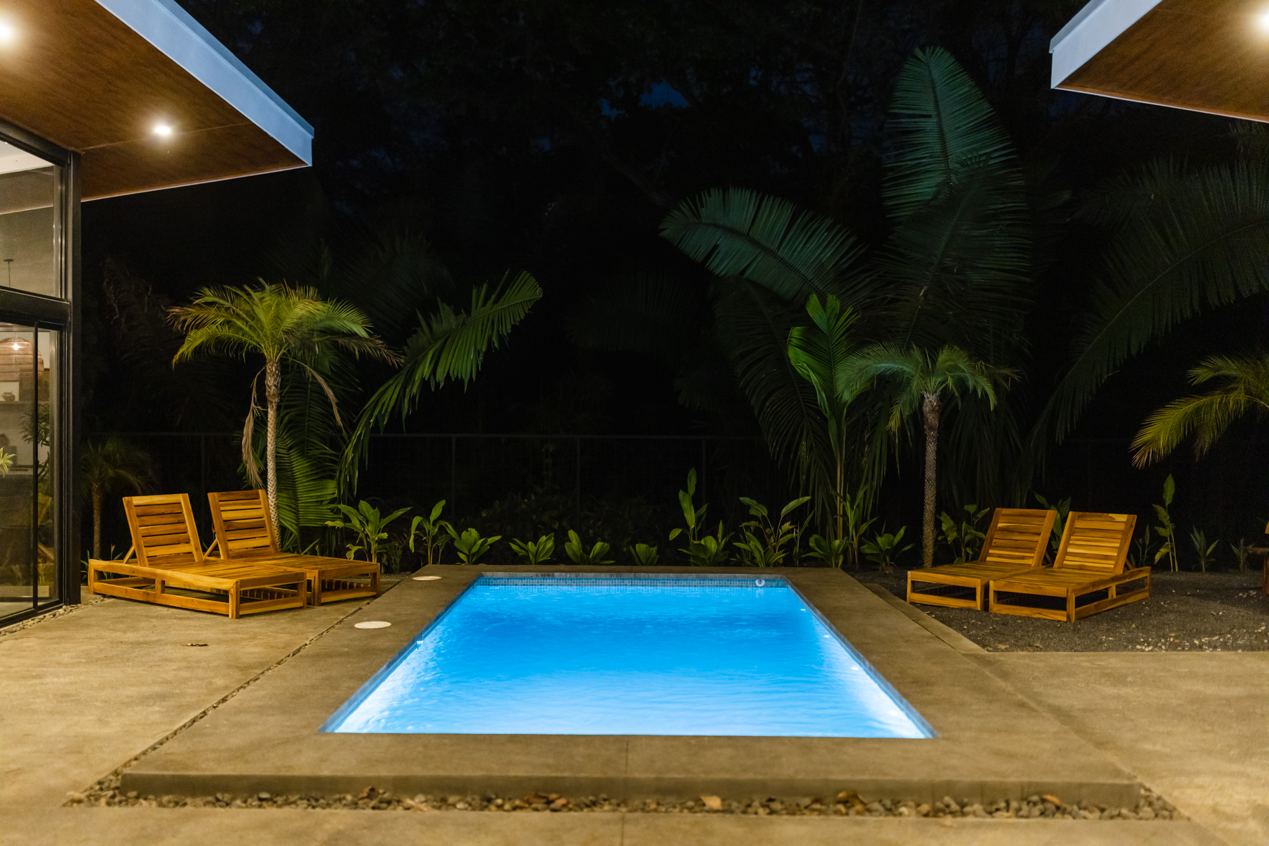 Glowing blue pool at night surrounded by palms and teak loungers